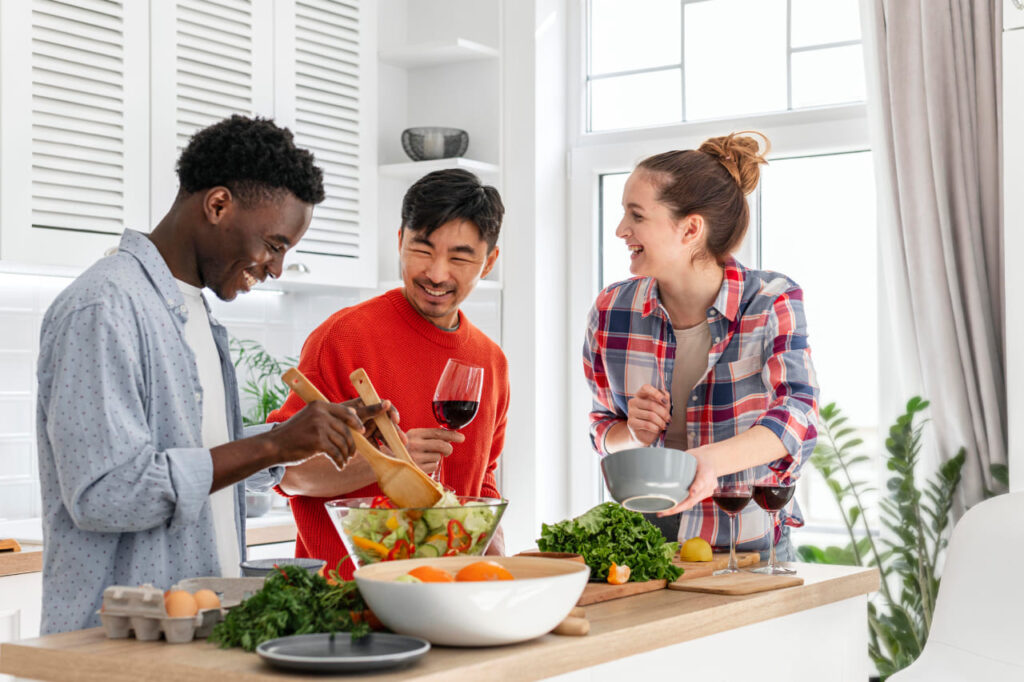 Amigos sorrindo na cozinha, preparando uma refeição com ingredientes frescos. Uma representação visual de relacionamentos saudáveis e união em torno da culinária.