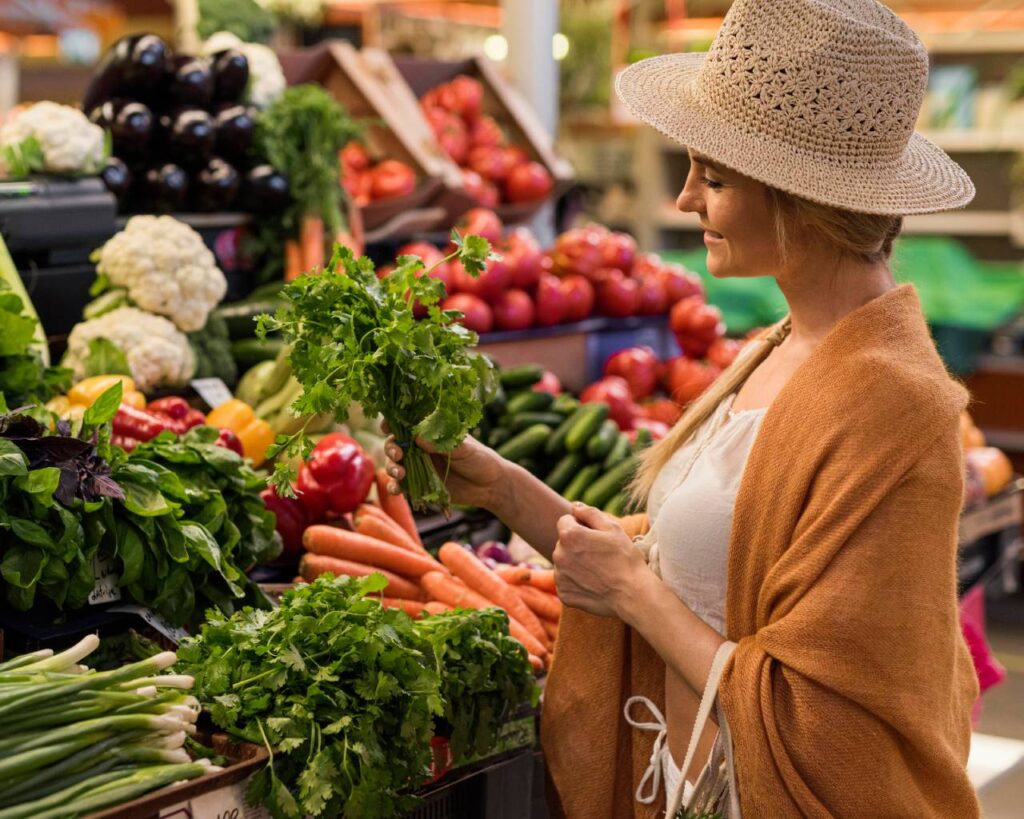 Mulher com chapéu escolhendo legumes frescos na feira, promovendo os Alimentos da Estação.