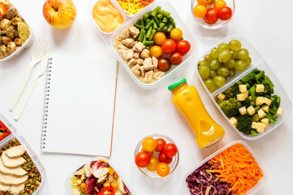 Imagem de marmitas saudáveis dispostas ao redor de um caderno aberto em uma página em branco. As marmitas exibem ingredientes frescos e variados, enquanto o caderno oferece a possibilidade de anotações e planejamento de refeições equilibradas. A imagem destaca a conexão entre alimentação saudável e organização, incentivando escolhas conscientes para um estilo de vida equilibrado.