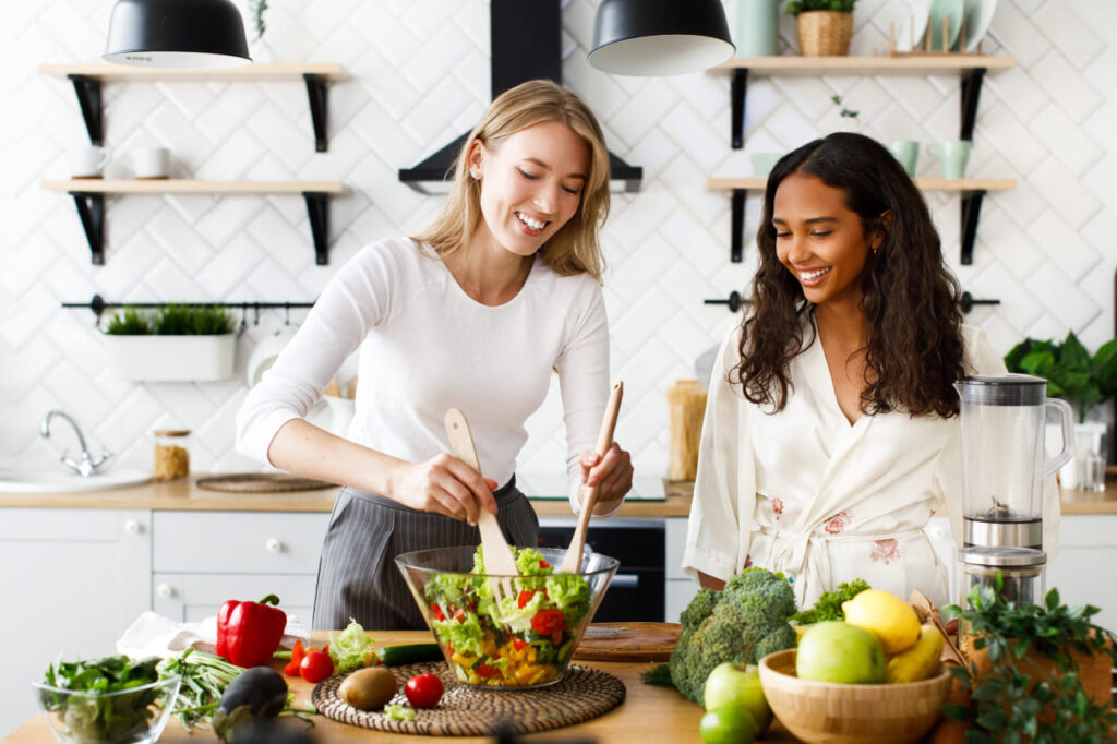 Alimentação equilibrada e saudável: Duas amigas sorrindo, preparando uma salada com ingredientes frescos.