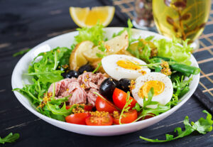 Salada saudável com alface, rúcula, tomate cereja, ovo cozido, legumes, atum e azeitona, acompanhado de uma bebida e laranja.