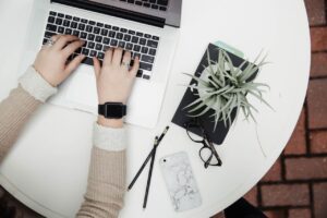 Mãos de uma mulher sobre um laptop em uma mesa de trabalho com itens de escritório ao redor.