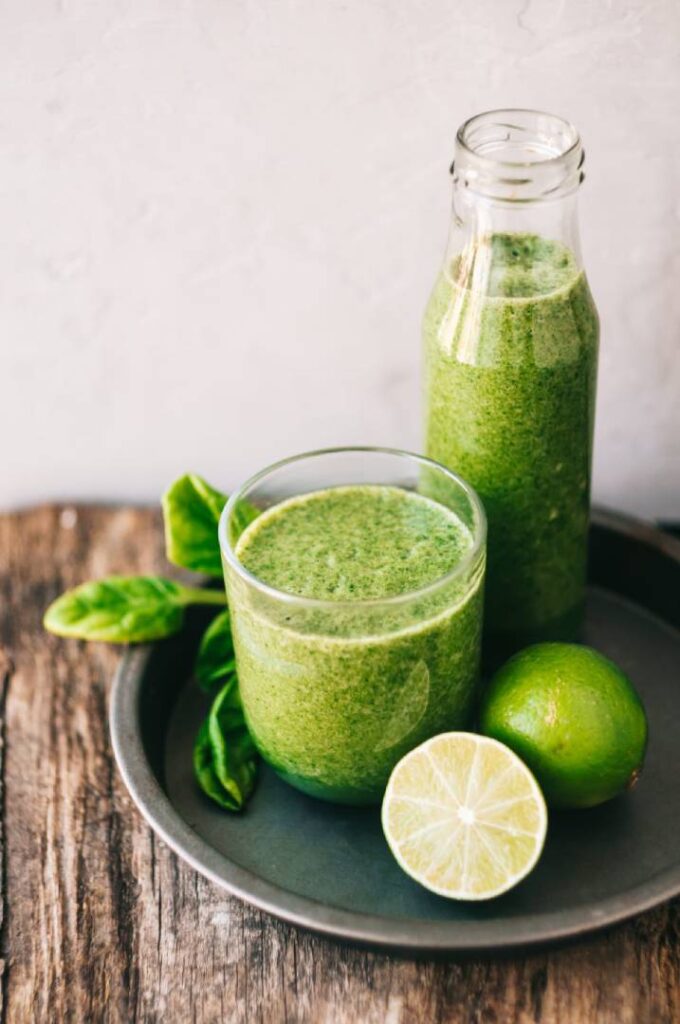 Suco Verde Emagrecedor em copo e garrafa de vidro, acompanhados por limão e folhas de espinafre em uma bandeja sobre mesa de madeira. Uma opção saudável para nutrir o corpo.