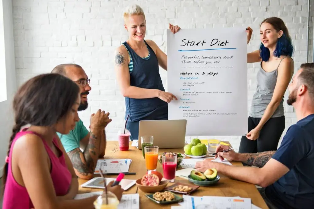 Grupo de colegas de trabalho em uma apresentação sobre alimentação saudável, com uma mesa cheia de alimentos nutritivos como maçã, romã, castanhas, abacate, salada, sucos e água, promovendo a alimentação consciente no trabalho.