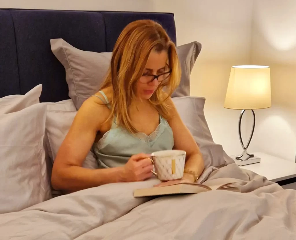 Imagem de uma mulher relaxando na cama com uma xícara de chá e um livro antes de dormir, demonstrando a preparação para uma rotina matinal saudável.