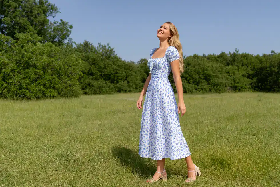 Mulher sorridente em um campo verde usando vestido floral azul e sandália, representando leveza, equilíbrio, autenticidade e bem-estar em um ambiente natural.