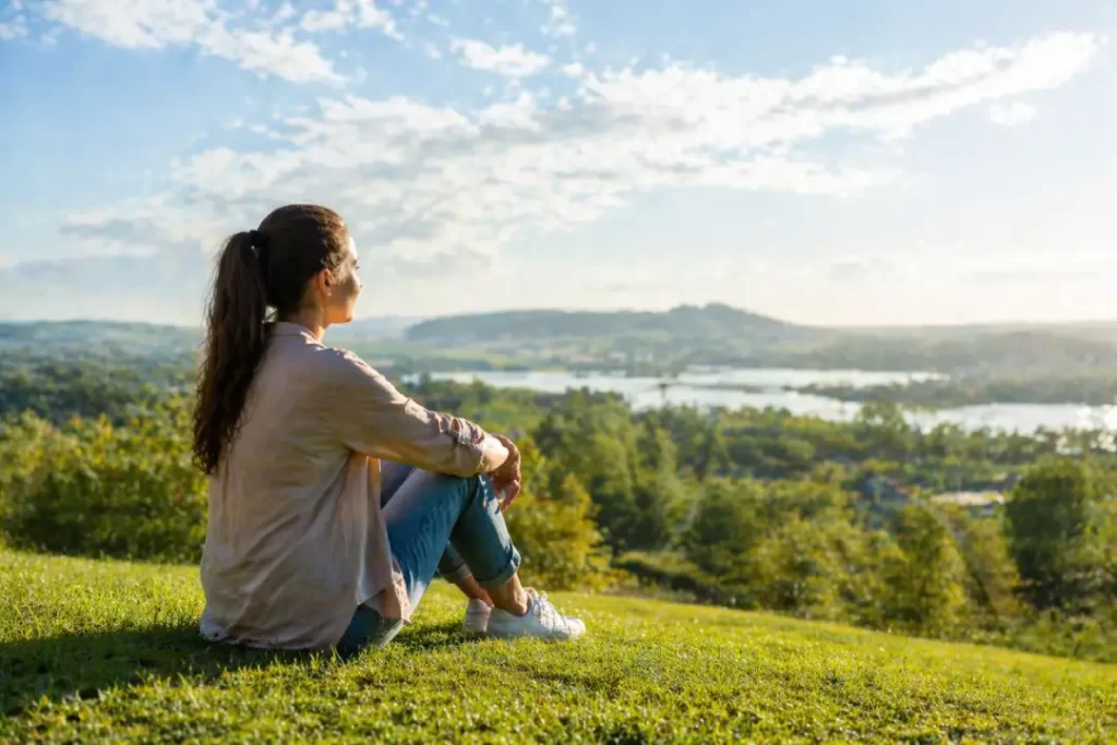 Mulher sentada na grama olhando o horizonte, representando natureza como terapia e momento de calma ao ar livre.